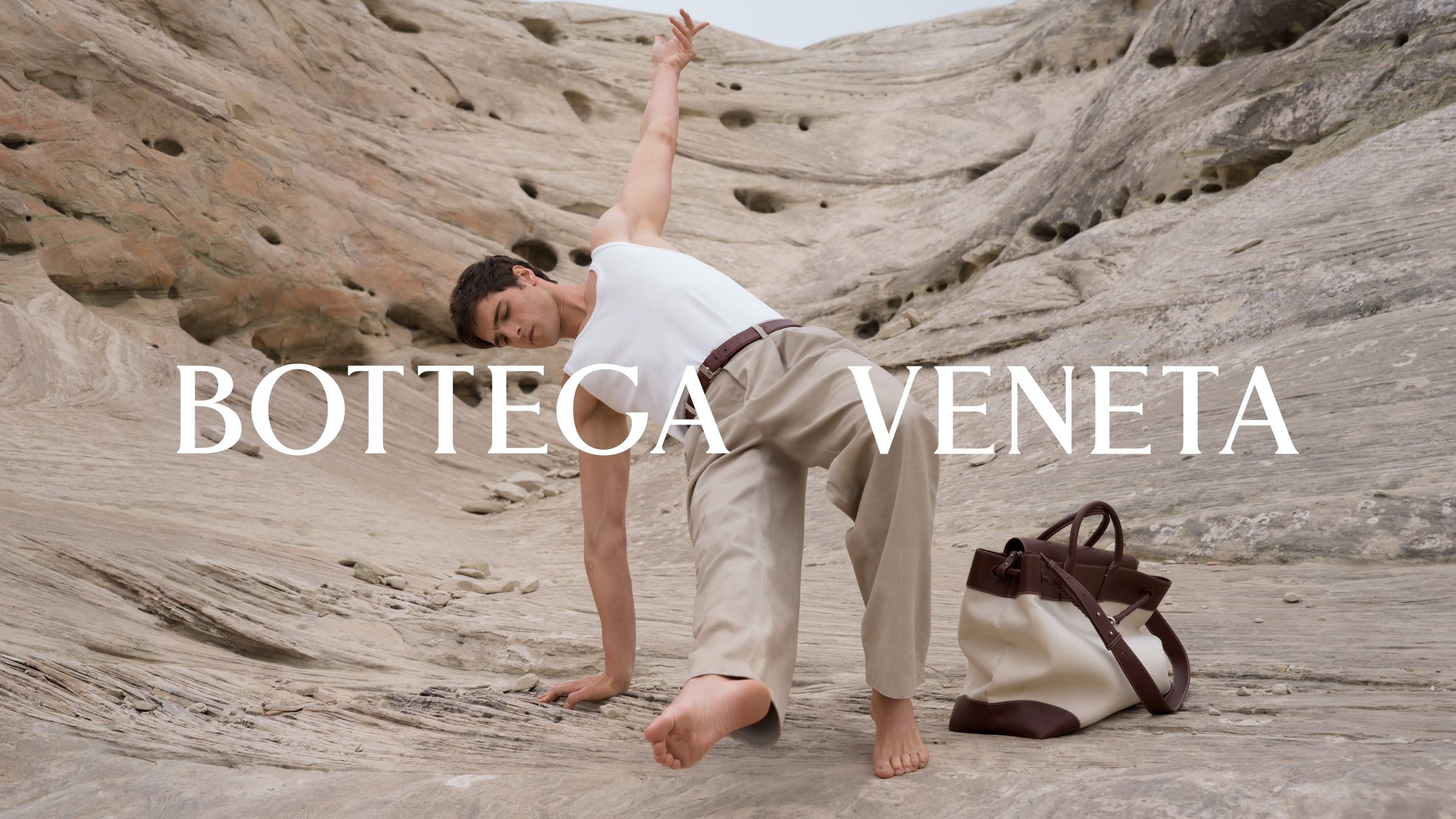 Model Jacob Elordi posing dynamically on eroded rocky terrain with a brown and white tote bag for Bottega Veneta