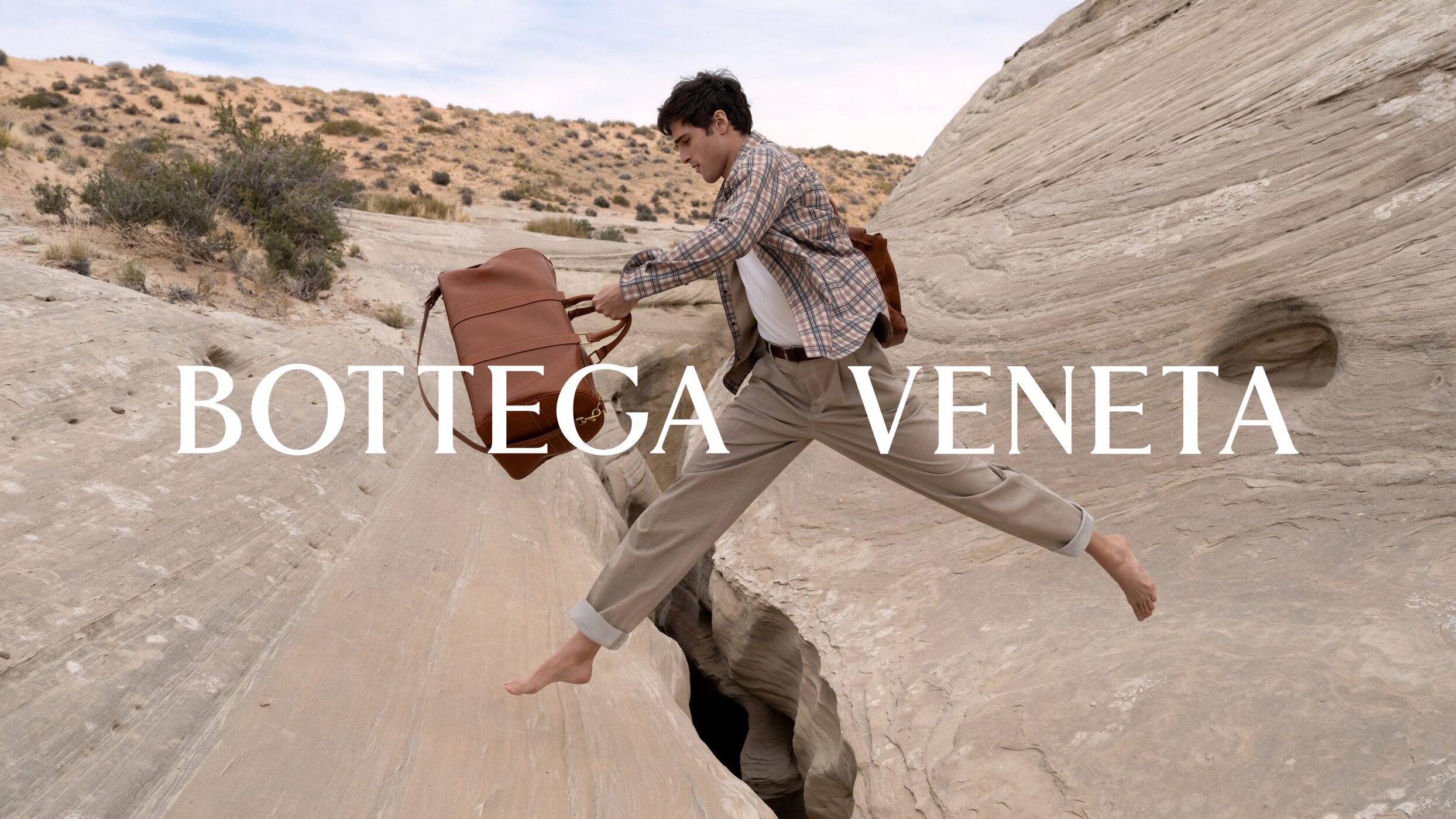 Man leaping barefoot over a rocky desert canyon holding a leather duffel bag