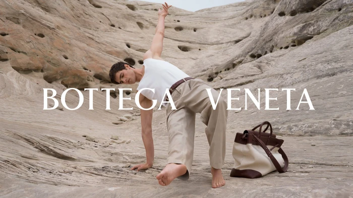 Model Jacob Elordi posing dynamically on eroded rocky terrain with a brown and white tote bag for Bottega Veneta