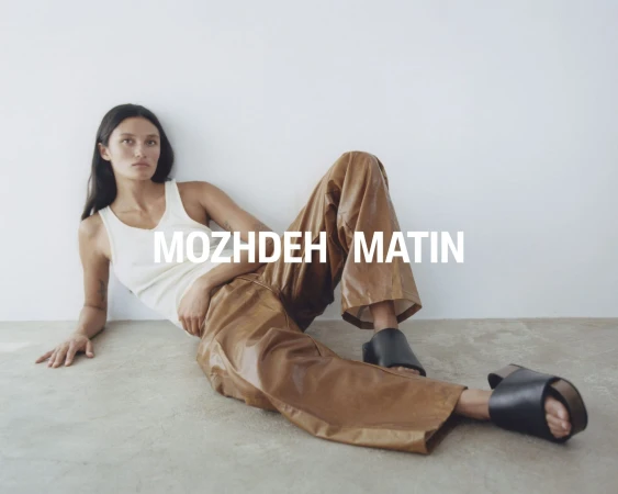 Model wearing a white tank top and brown trousers reclining on a floor against a plain wall