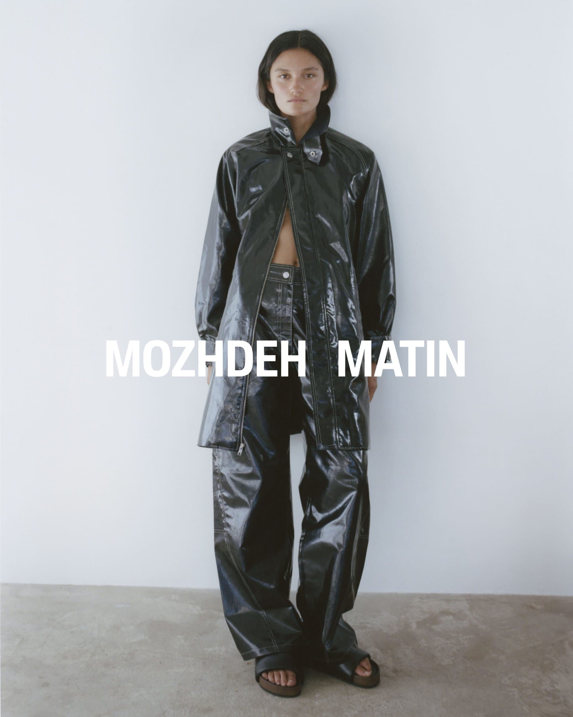 Model wearing a glossy black leather coat and trousers standing against a plain white studio background.