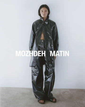 Model wearing a glossy black leather coat and trousers standing against a plain white studio background.