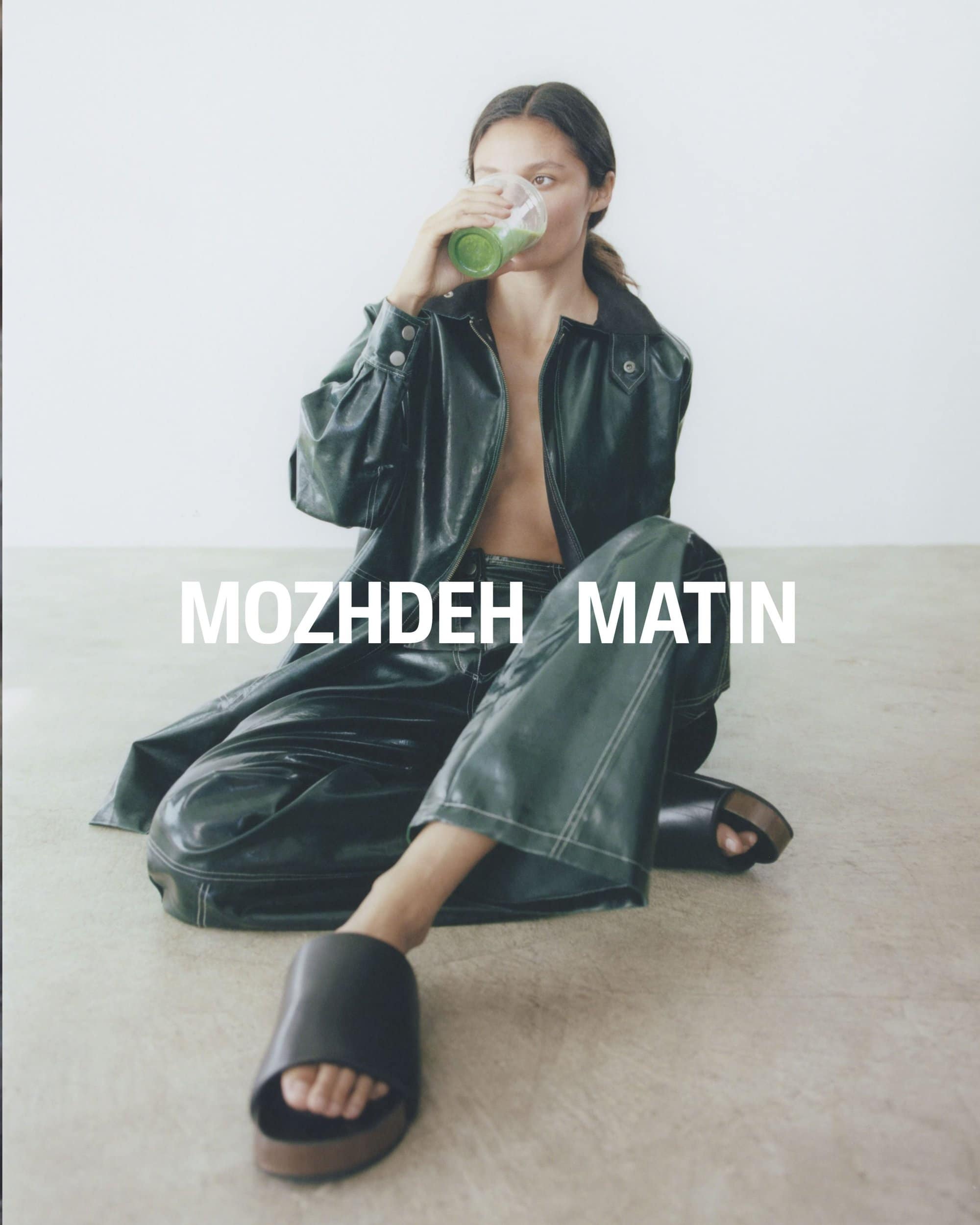 Model wearing a dark green leather jacket and trousers seated on the floor drinking from a cup against a neutral background.