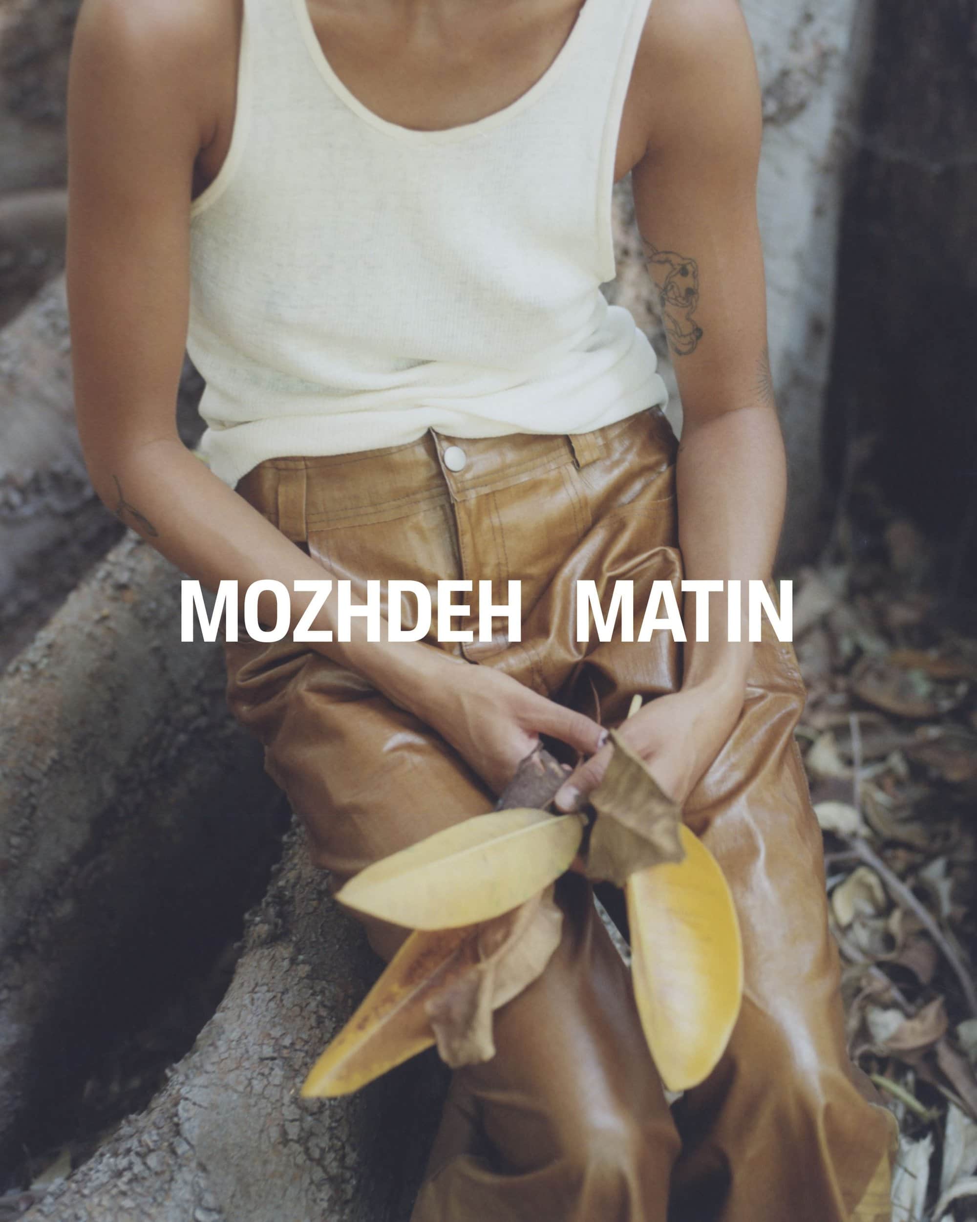 Model wearing a white tank top and brown leather trousers holding fallen leaves against a textured woodland ground