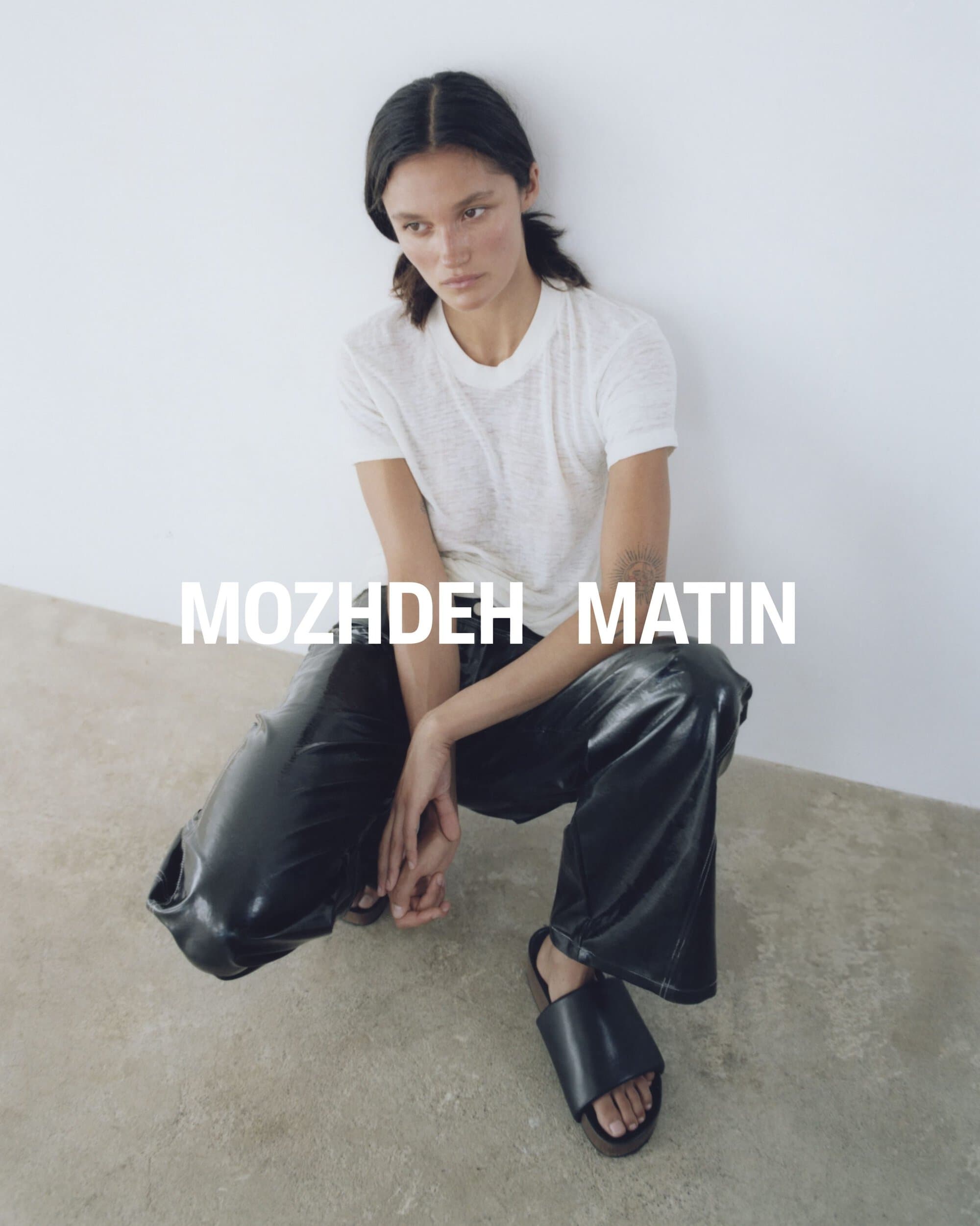Model crouching wearing a white t-shirt, black leather trousers, and black sandals against a white studio wall