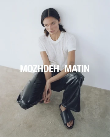 Model crouching wearing a white t-shirt, black leather trousers, and black sandals against a white studio wall