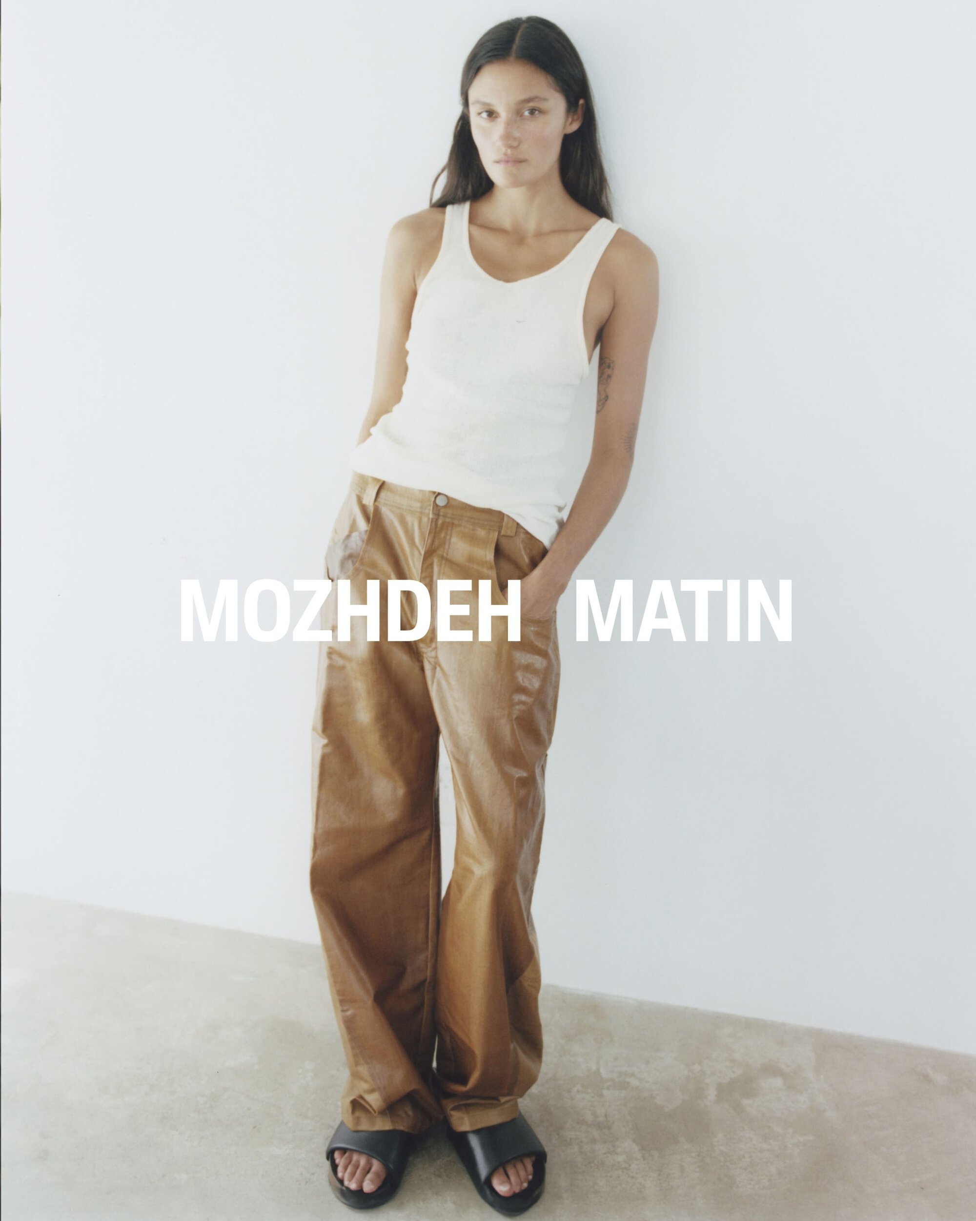 Model wearing a white tank top and brown trousers leaning against a white studio wall