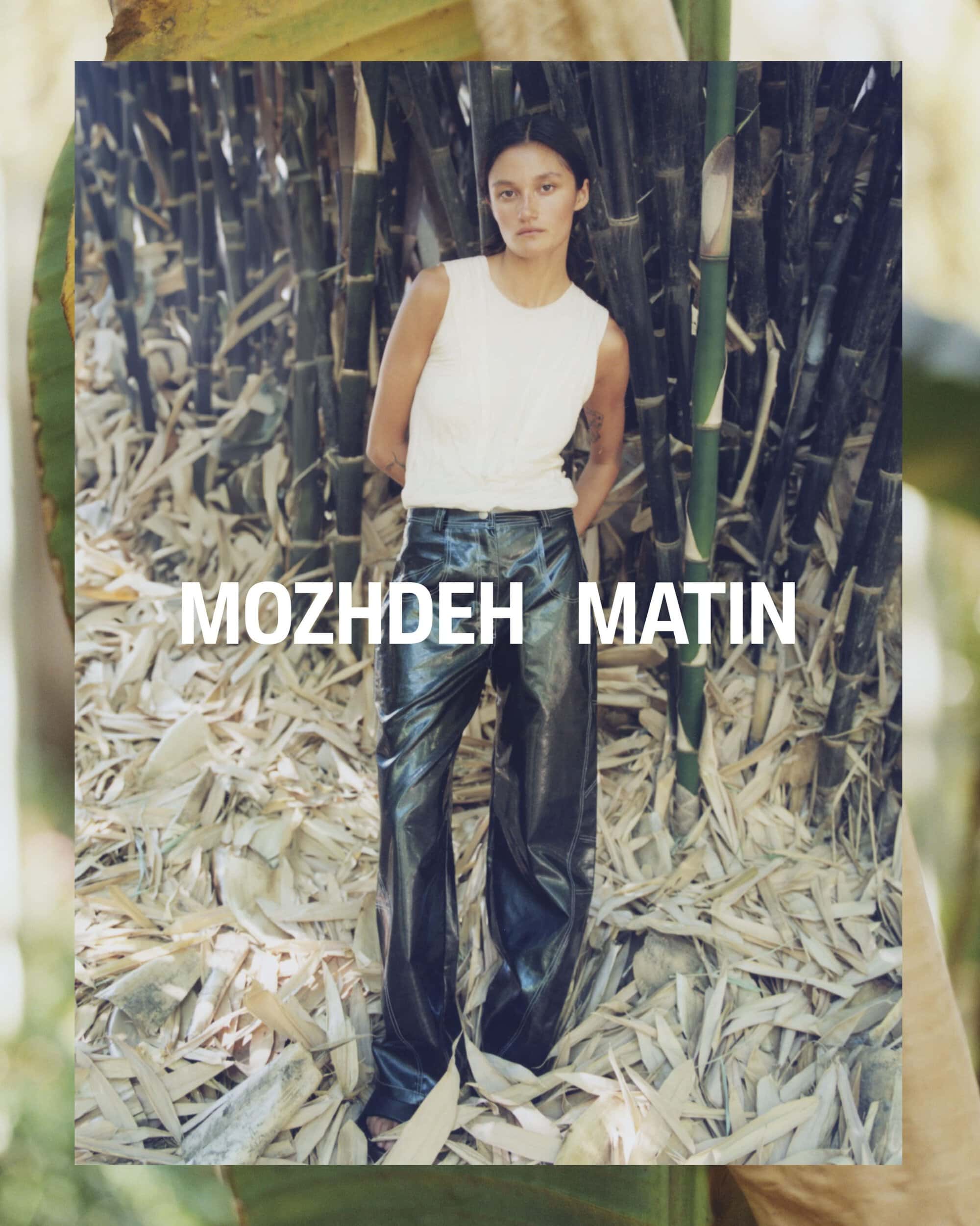 Model standing in a white sleeveless top and dark trousers against a dense background of vertical bamboo stalks.