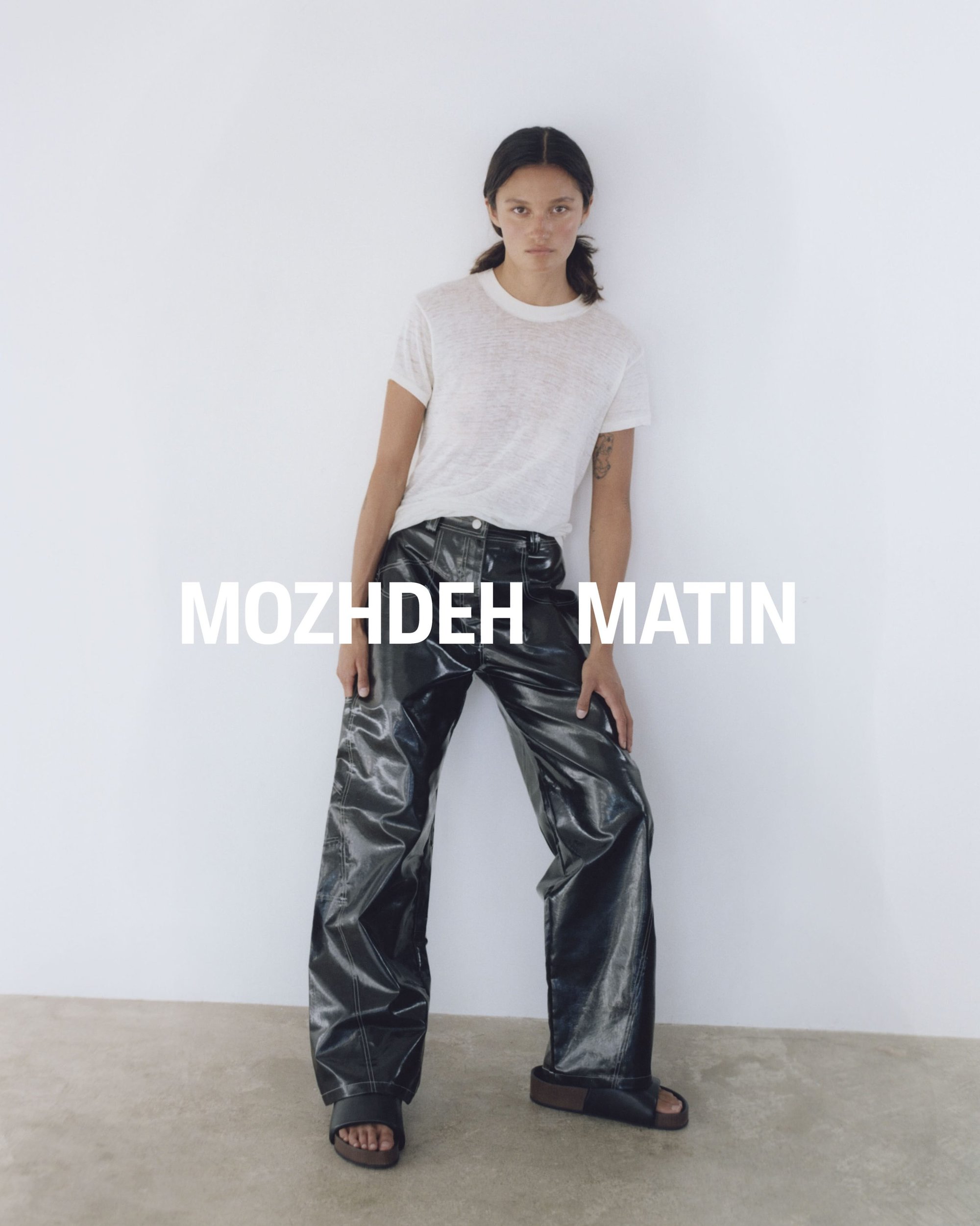 Model wearing a white t-shirt and black glossy trousers standing against a plain white studio wall for Mozhdeh Matin.