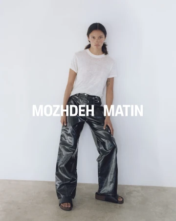 Model wearing a white t-shirt and black glossy trousers standing against a plain white studio wall for Mozhdeh Matin.