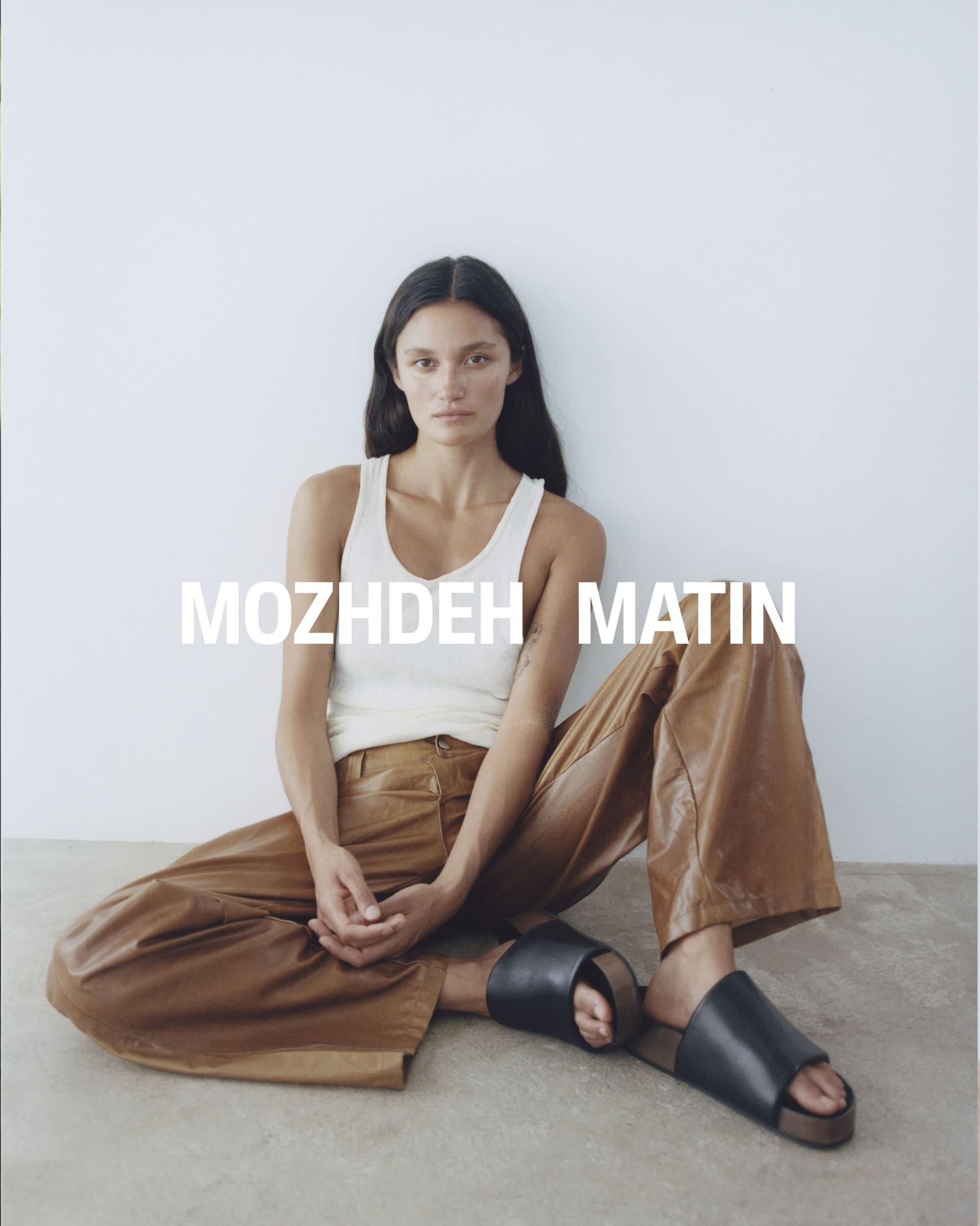 Model seated on the floor wearing a white tank top and brown trousers against a plain white background