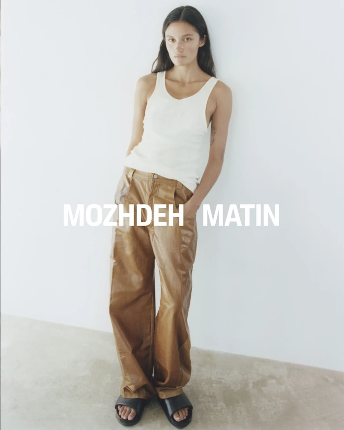 Model wearing a white tank top and brown trousers leaning against a white studio wall
