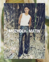 Model standing in a white sleeveless top and dark trousers against a dense background of vertical bamboo stalks.