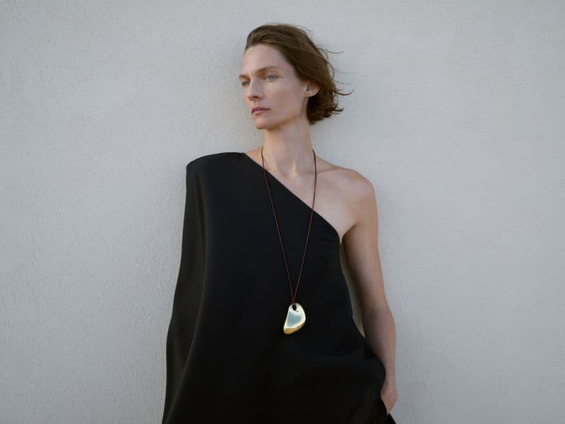 Model Karolin Wolter wearing a black one-shoulder dress and gold pendant necklace against a light background