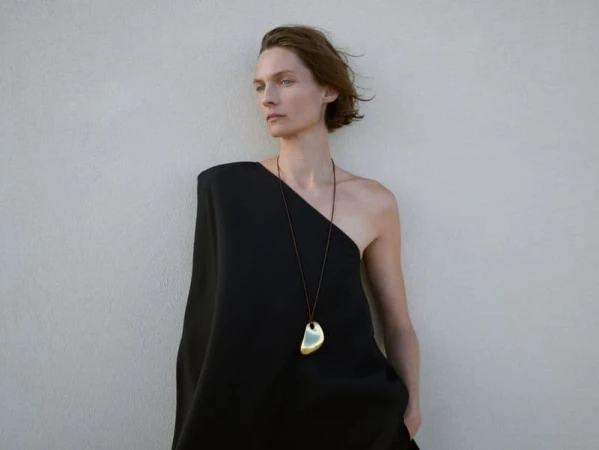 Model Karolin Wolter wearing a black one-shoulder dress and gold pendant necklace against a light background
