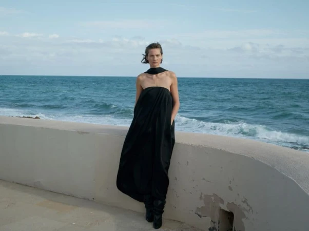 Model in a long black strapless dress leaning against a low concrete sea wall with the ocean in the background.