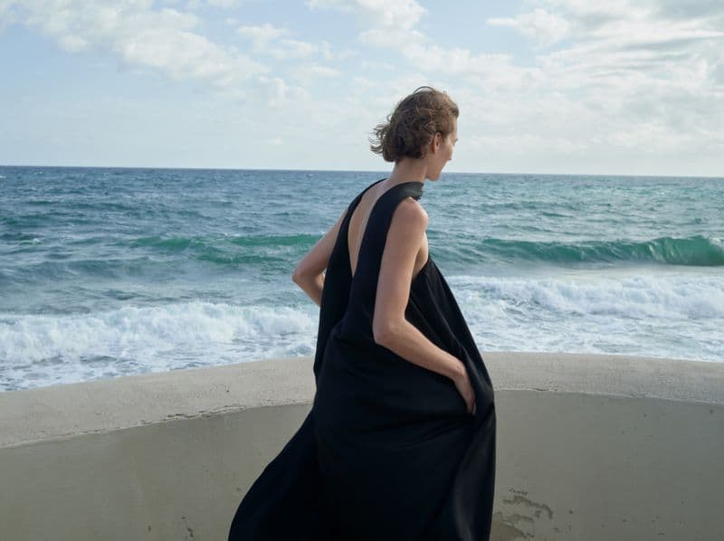 Model wearing a black sleeveless dress standing on a coastal wall overlooking the sea for Alexandre Vauthier spring 2025 ad campaign.