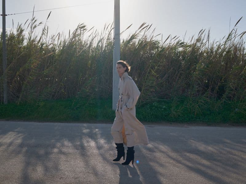 Model wearing a long beige trench coat and tall black boots standing on an outdoor asphalt road with tall green reeds in background