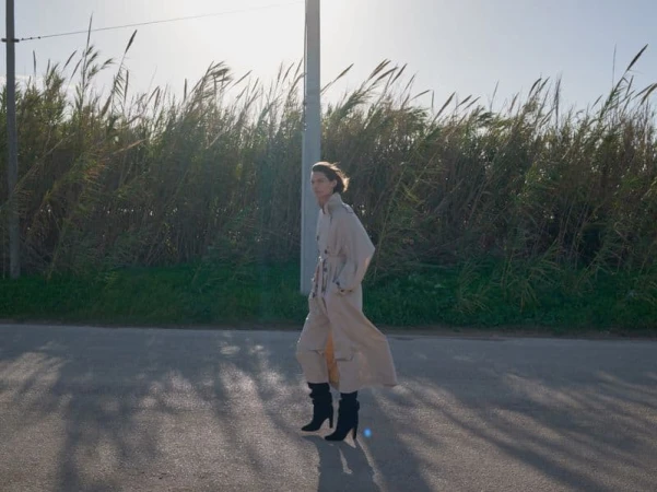 Model wearing a long beige trench coat and tall black boots standing on an outdoor asphalt road with tall green reeds in background