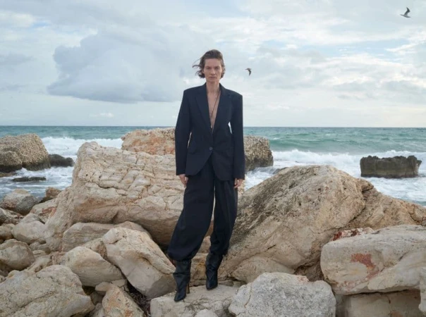 Model standing on rocky coastal shore wearing a dark blazer and trousers with the ocean behind her