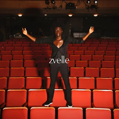 A Black man in a black shirt and trousers standing with arms outstretched on red theatre seats.