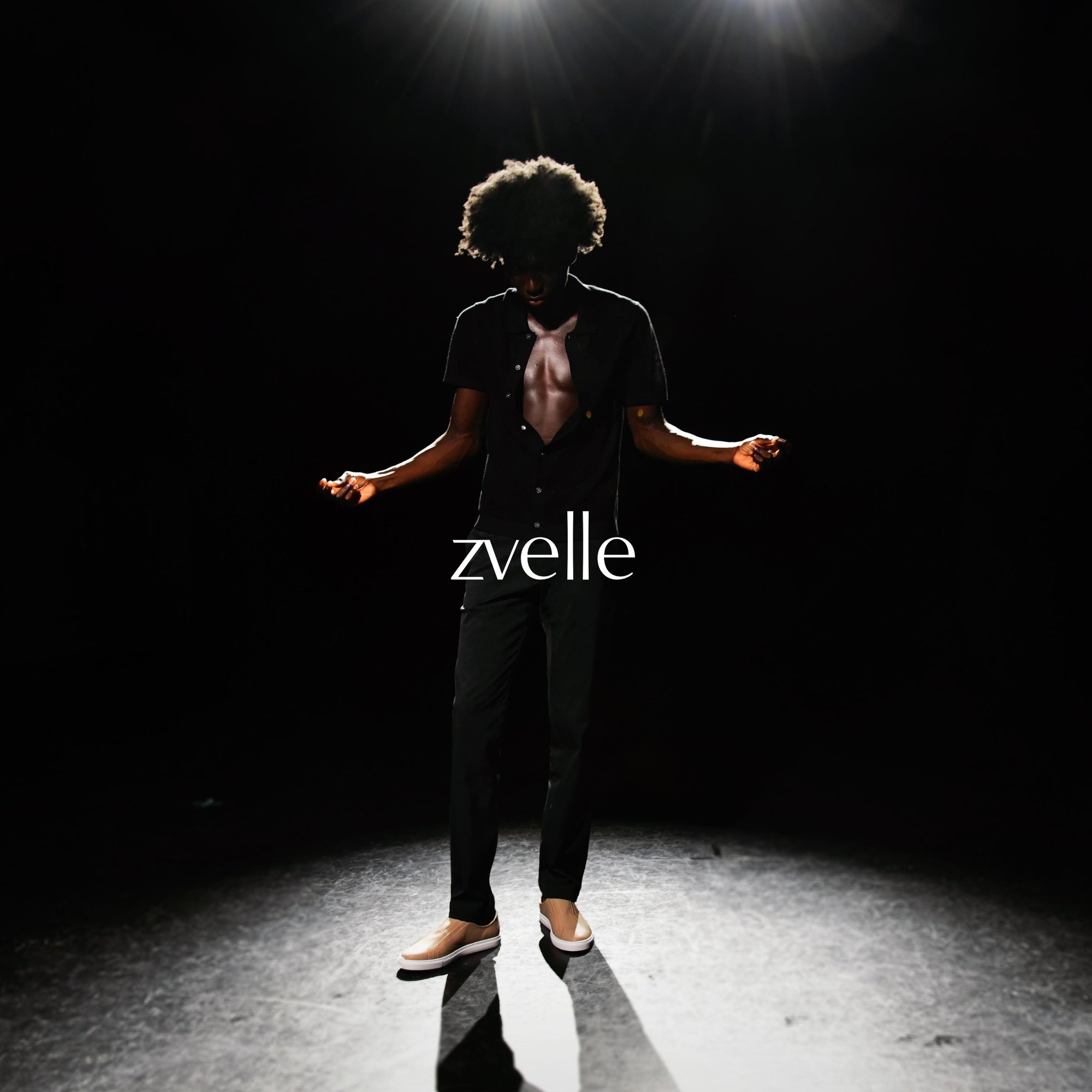 A man standing under overhead spotlight wearing a dark unbuttoned shirt and pants against a black background with brand text zvelle