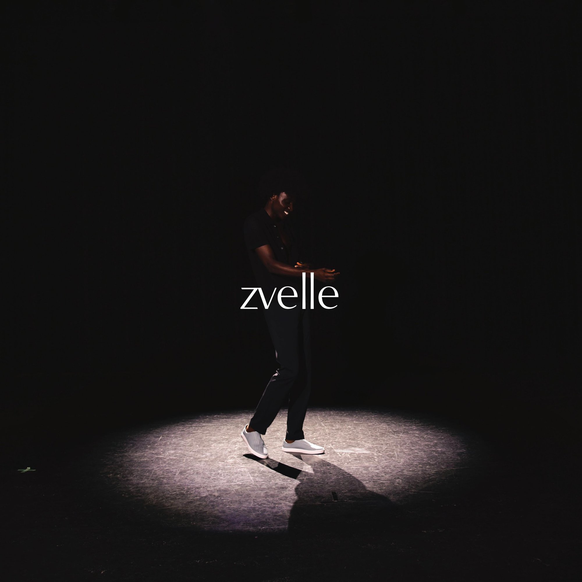 Model standing in a spotlight wearing dark clothing and white sneakers with the zvelle brand name displayed across the center