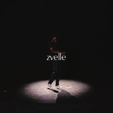 Model standing in a spotlight wearing dark clothing and white sneakers with the zvelle brand name displayed across the center