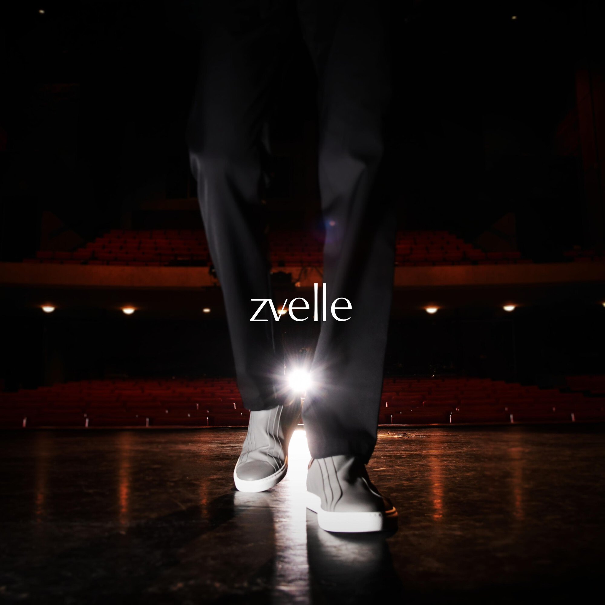 Close-up shot of a model standing on a stage wearing dark trousers and grey sneakers with white soles for Zvelle Fall 2024 campaign.