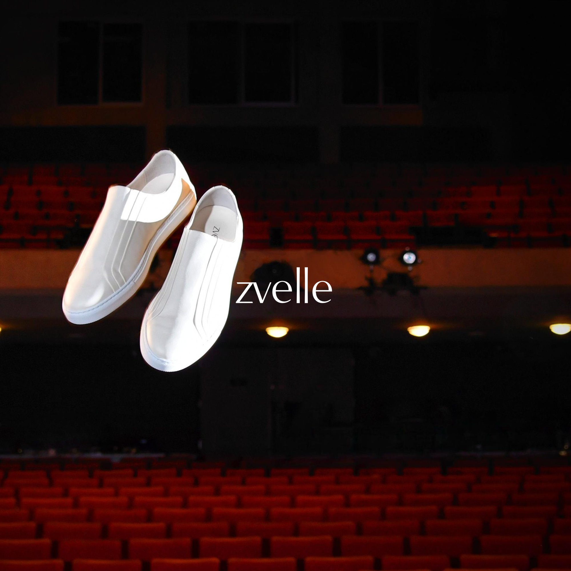 Two white slip-on sneakers floating against a dark auditorium with rows of red theatre seats.