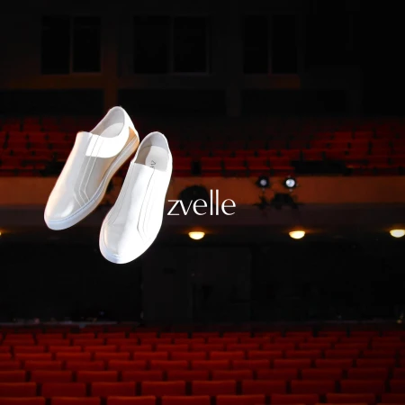 Two white slip-on sneakers floating against a dark auditorium with rows of red theatre seats.