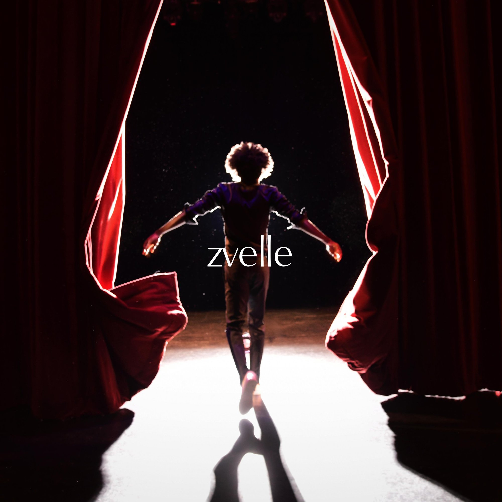 A person viewed from behind with arms outstretched standing before parted red theatre curtains under a bright spotlight