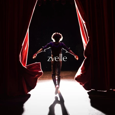 A person viewed from behind with arms outstretched standing before parted red theatre curtains under a bright spotlight