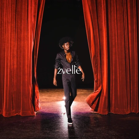 Model walking between heavy red velvet theatre curtains on a wooden stage with dramatic lighting for Zvelle Fall 2024 campaign