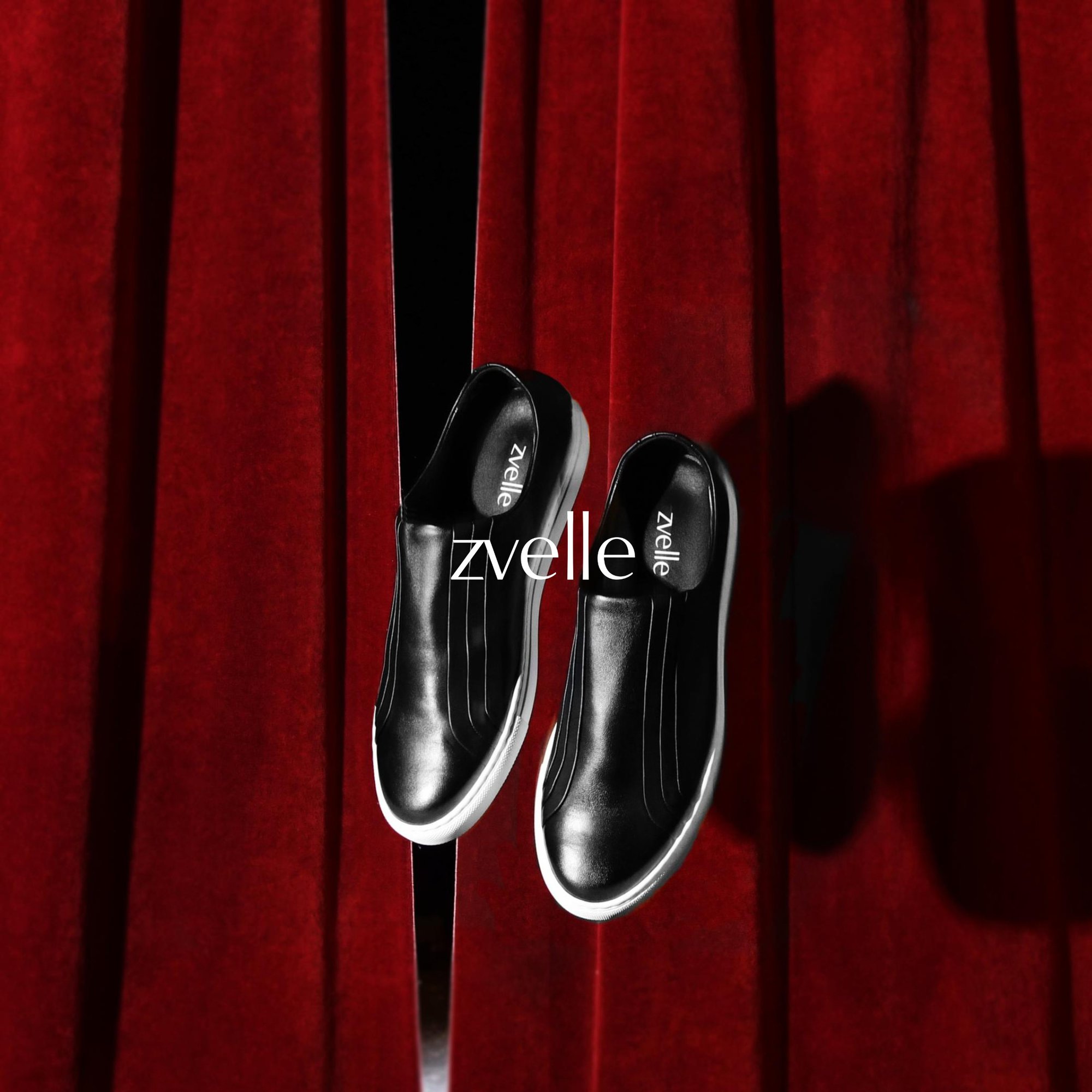 Two black slip-on shoes with white soles suspended against red velvet curtains for Zvelle Fall 2024 campaign