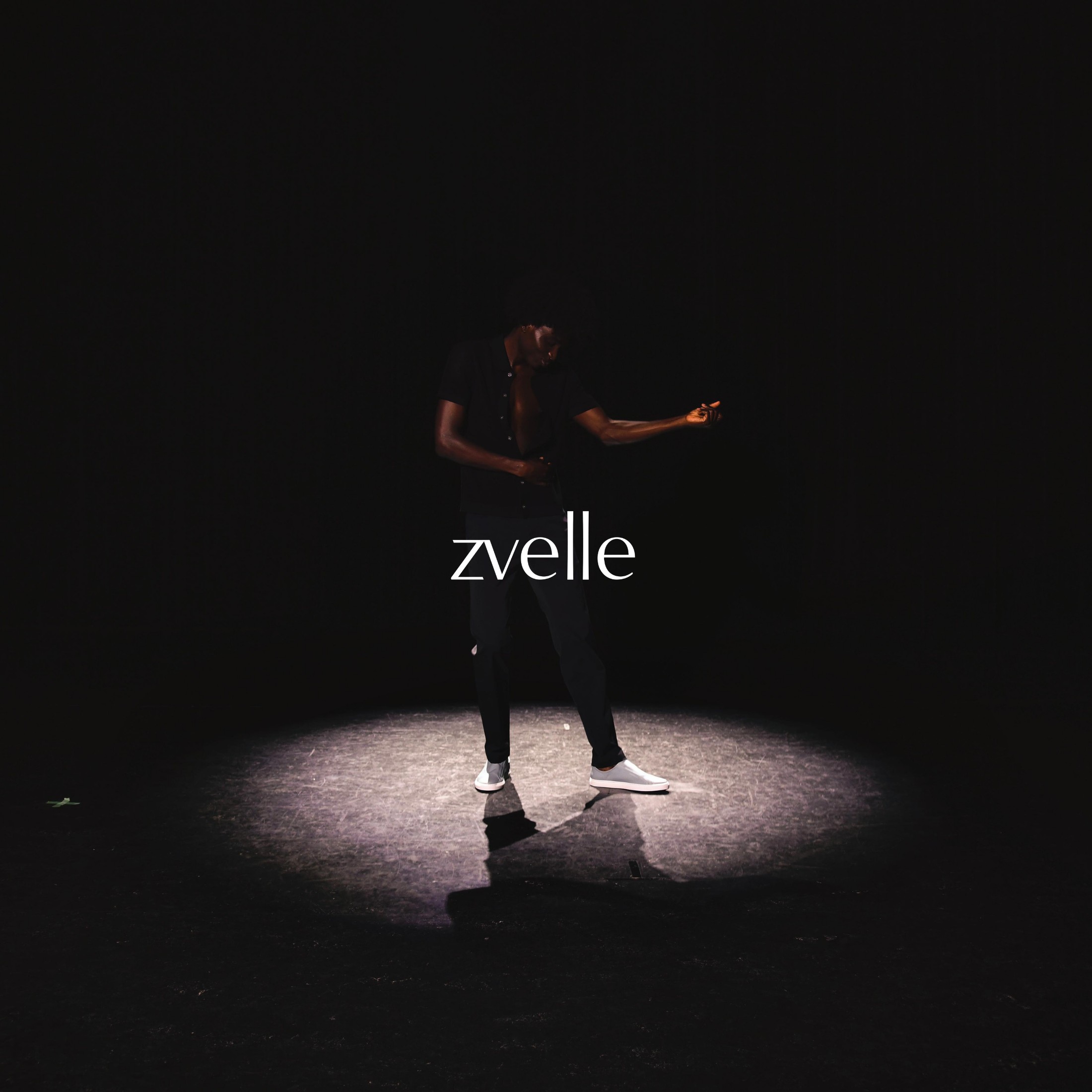 A man standing in a dramatic spotlight on a dark background wearing dark clothing and light-colored shoes, featuring the zvelle logo.