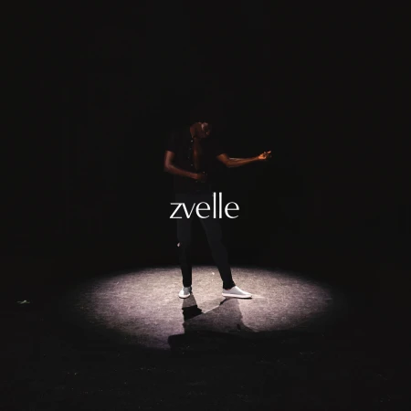 A man standing in a dramatic spotlight on a dark background wearing dark clothing and light-colored shoes, featuring the zvelle logo.