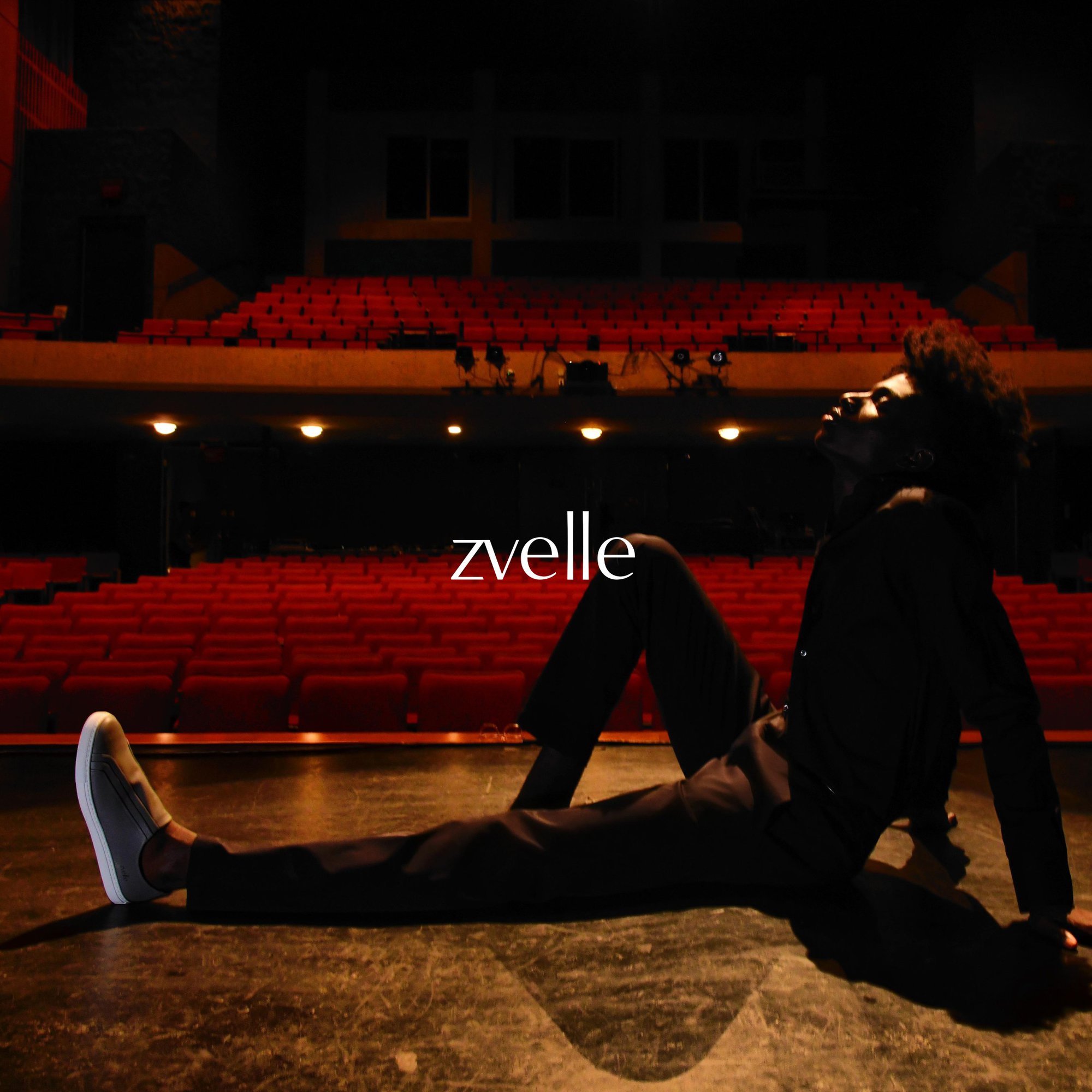 Model seated on a darkened stage with rows of red theatre seats in the background for Zvelle Fall 2024 ad campaign.