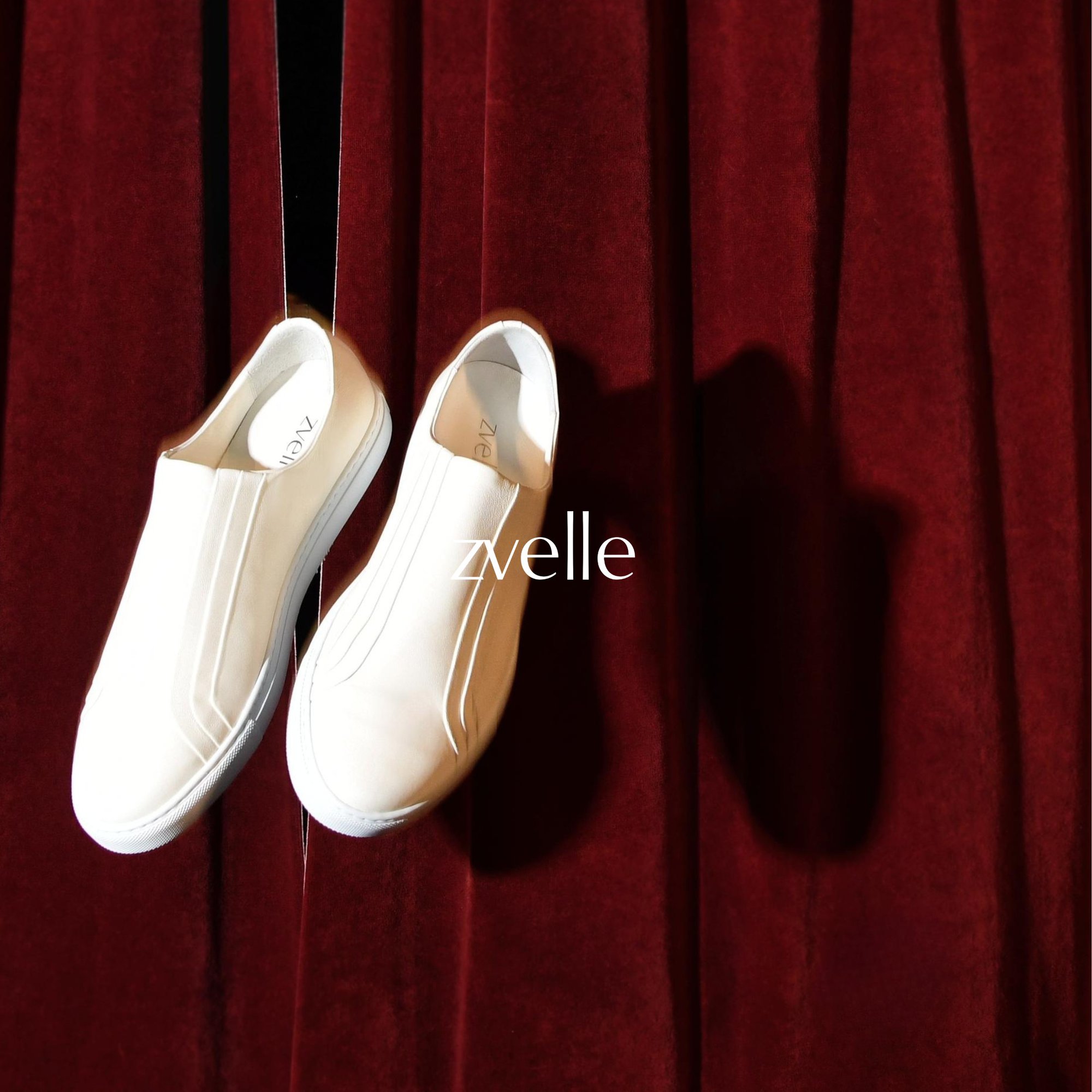 Two white slip-on sneakers hanging against a rich red velvet curtain background for Zvelle