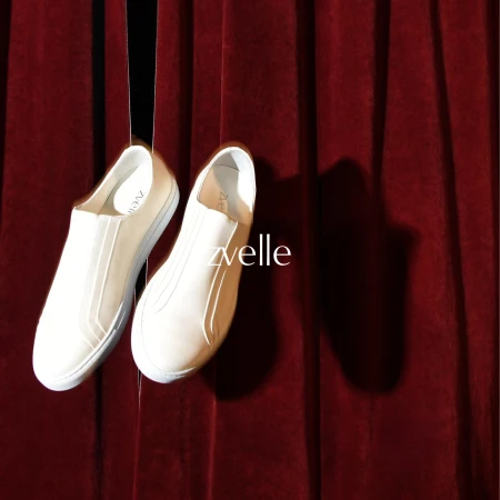 Two white slip-on sneakers hanging against a rich red velvet curtain background for Zvelle