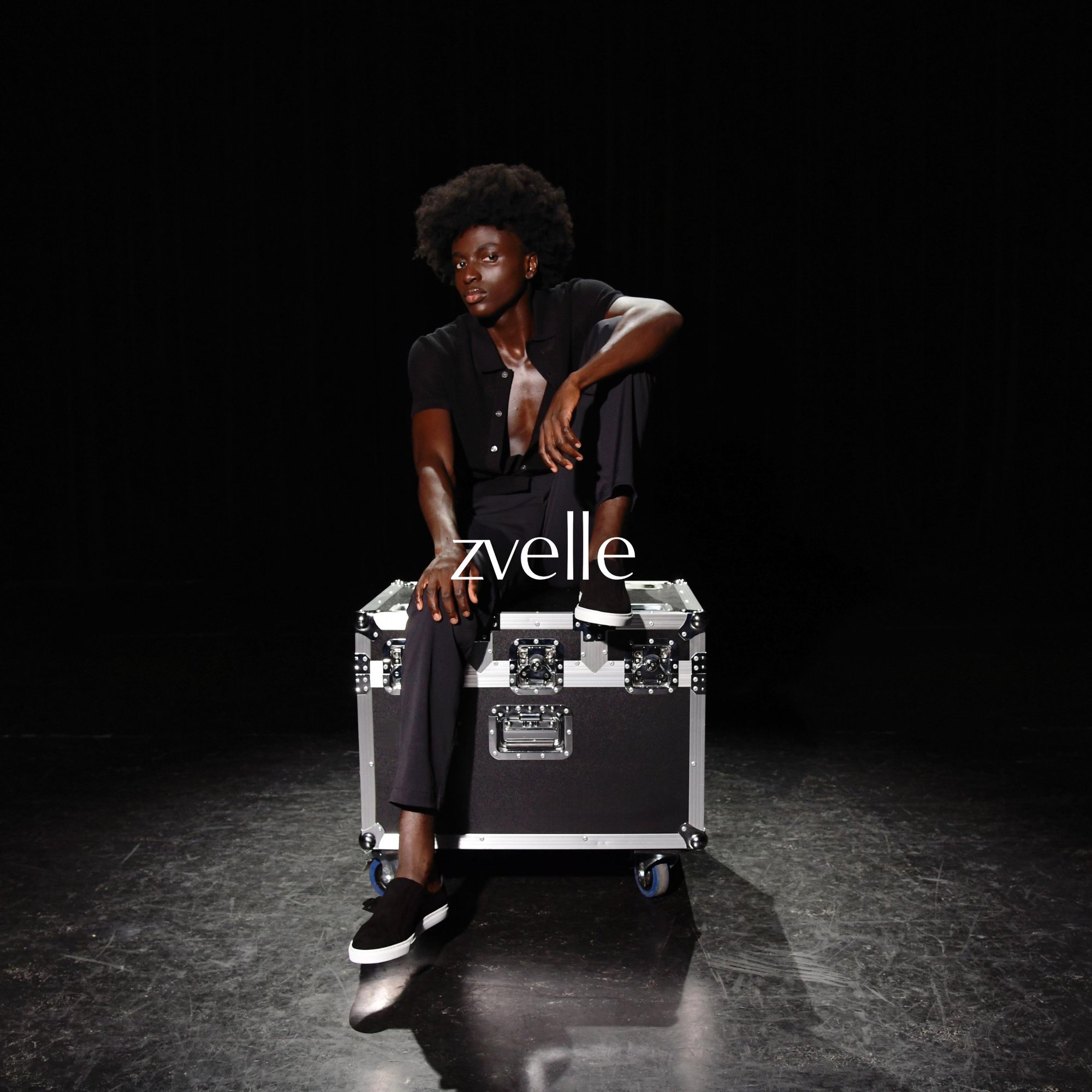 Black male model in dark clothing seated on a wheeled flight case against a black background