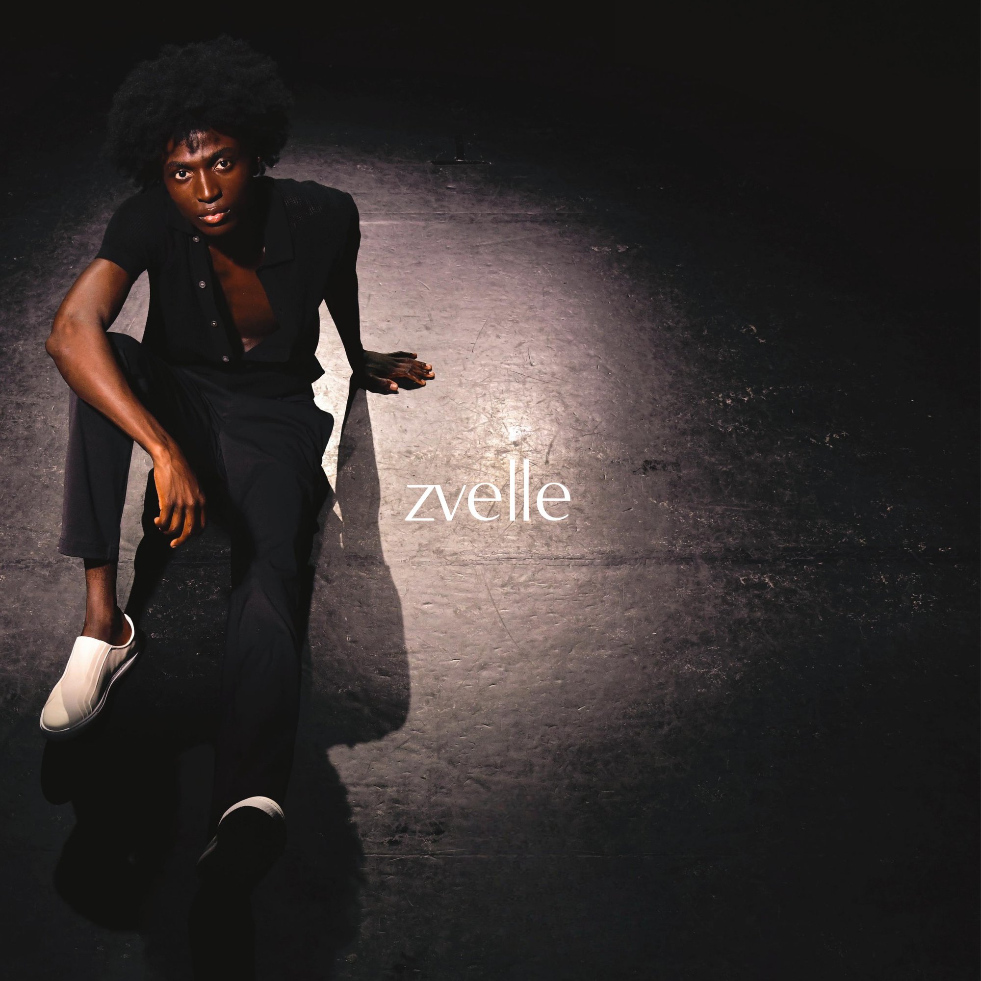Model seated on a dark floor in a spotlight wearing a black shirt and white slip-on shoes for Zvelle Fall 2024 campaign.