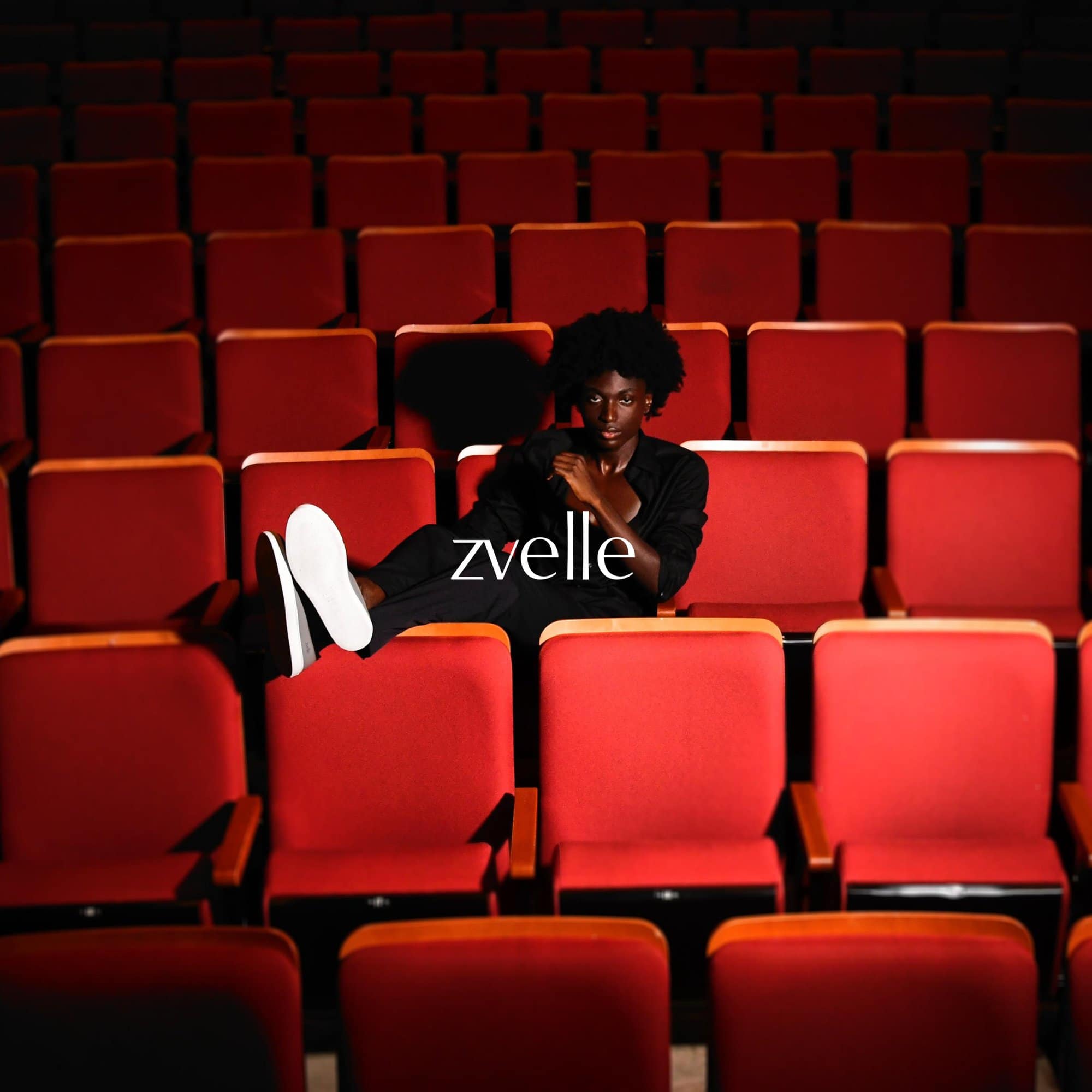 Model reclining across red theatre seats wearing black clothing and white sneakers for Zvelle