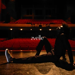 Model seated on a dimly lit theater stage wearing dark attire and grey sneakers with red rows of seats in the background