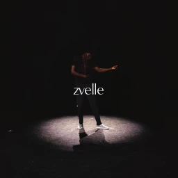 A man standing in a dramatic overhead spotlight on a dark background, wearing dark clothing and white shoes.