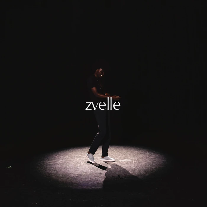 Model standing in a spotlight against a dark background wearing dark clothing and white slip-on sneakers for Zvelle fall 2024 ad campaign.