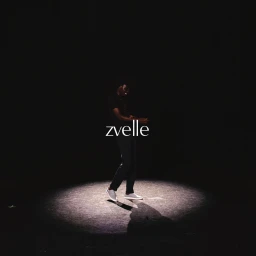 Model standing in a spotlight against a dark background wearing dark clothing and white slip-on sneakers for Zvelle fall 2024 ad campaign.
