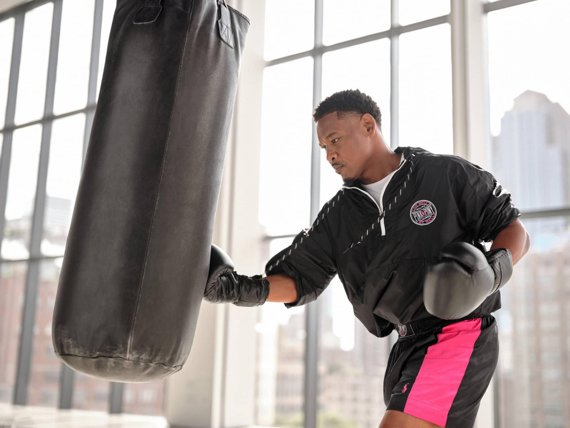 A man wearing a black jacket and pink-trimmed shorts punching a heavy black punching bag inside a studio.