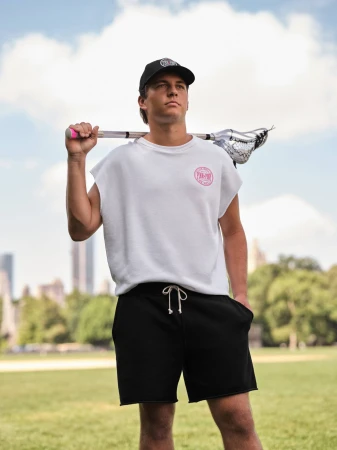 Young man wearing a white sleeveless shirt and black shorts holding a lacrosse stick outdoors against a park background