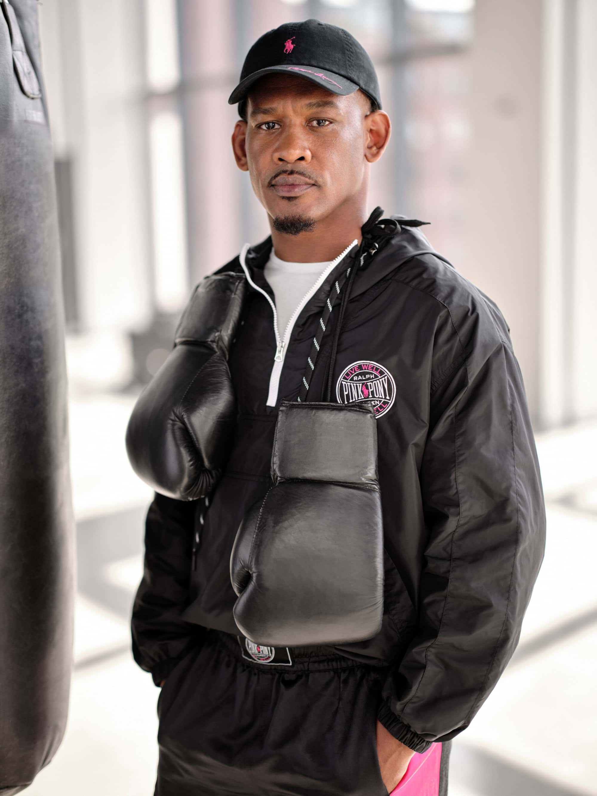 Man wearing a black windbreaker jacket and cap with boxing gloves draped around his neck in a bright gym setting